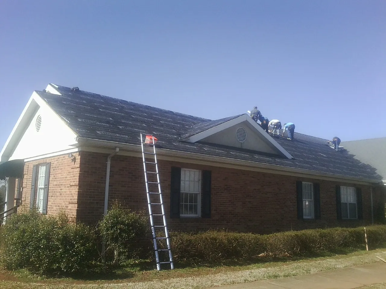 Crew tearing shingles off a brick ranch roof with a ladder set — Metal Roof Repair work by our crew in East Moline