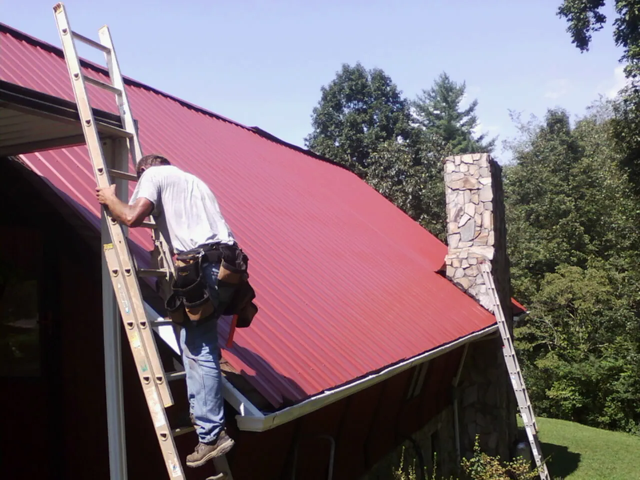 Roofer on a ladder beside a red metal roof and stone chimney — Soffit & Fascia Repair by Roofing Brothers in East Moline
